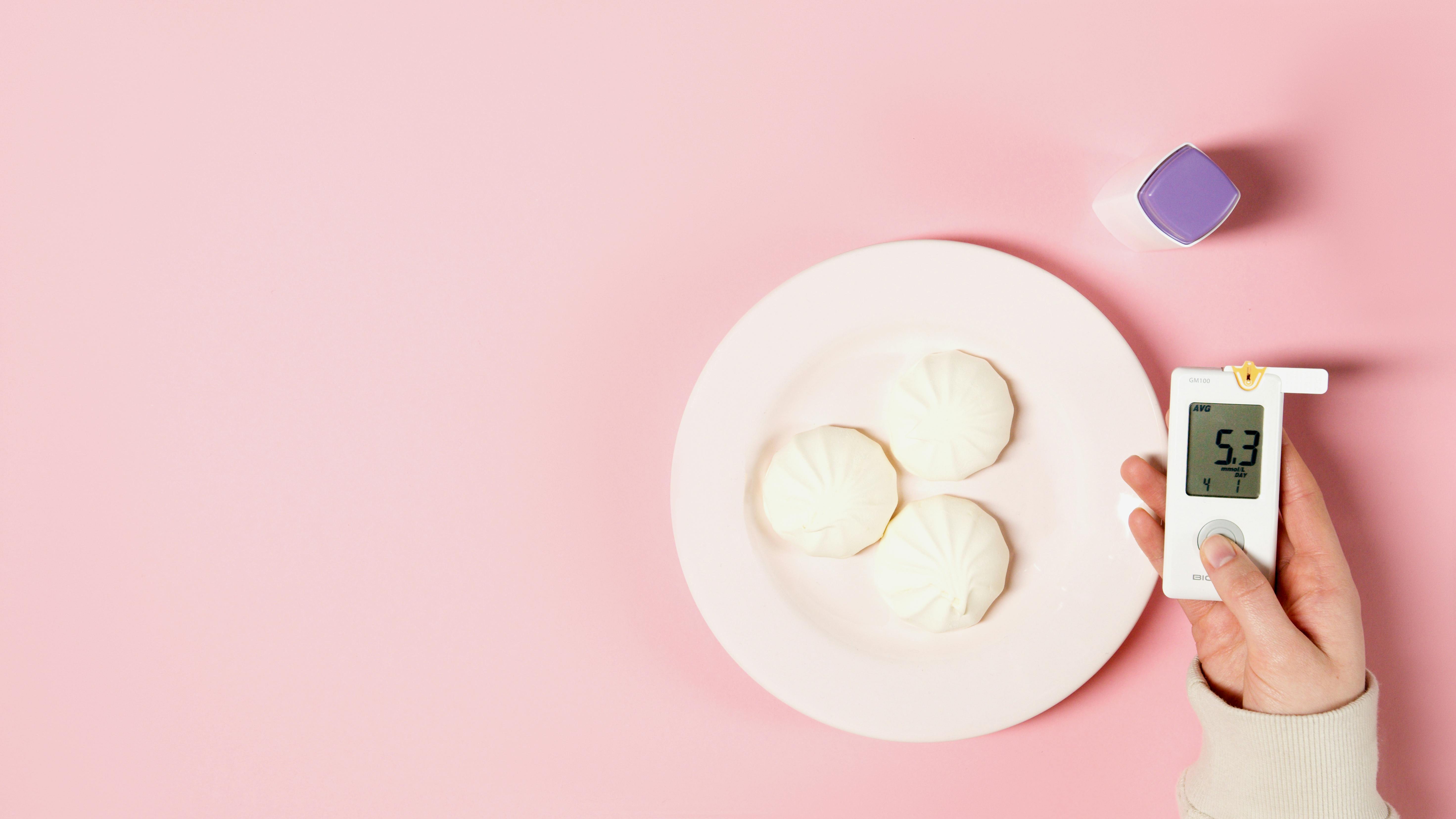 Flat lay of glucose meter and white sweets on a pink background, symbolizing diabetes awareness.