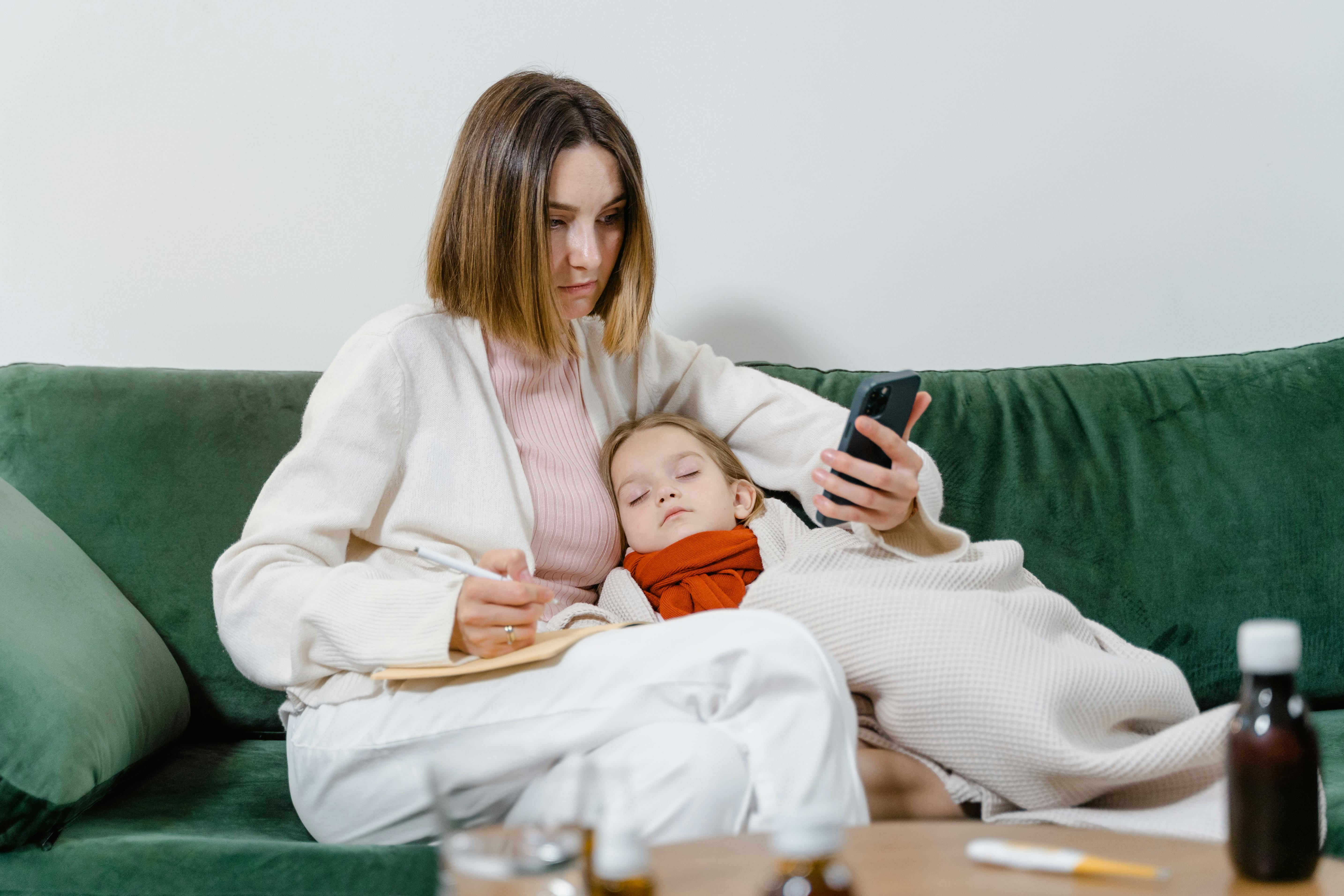 Woman researches symptoms on phone while comforting sleeping child on couch.